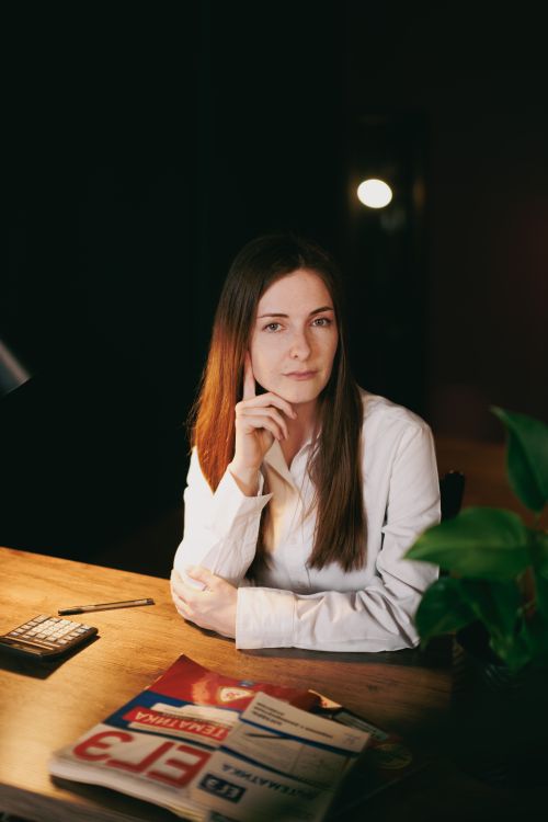 Free Young woman in a white shirt sits at a desk with magazines and calculator, exuding professionalism. Stock Photo