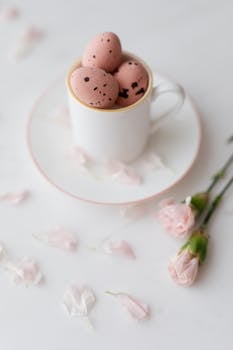 A stylish still life of pink eggs in a cup surrounded by delicate pink flowers on a white background.
