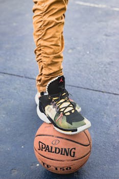 Crop unrecognizable male in trendy sneakers stepping on basketball ball while standing on sports ground