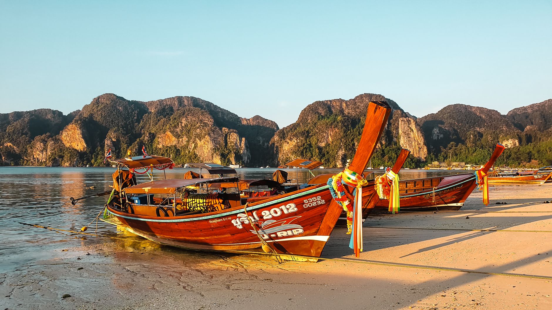 Explore the vibrant longtail boats on a tranquil Thai beach during sunset, surrounded by stunning karst formations.
