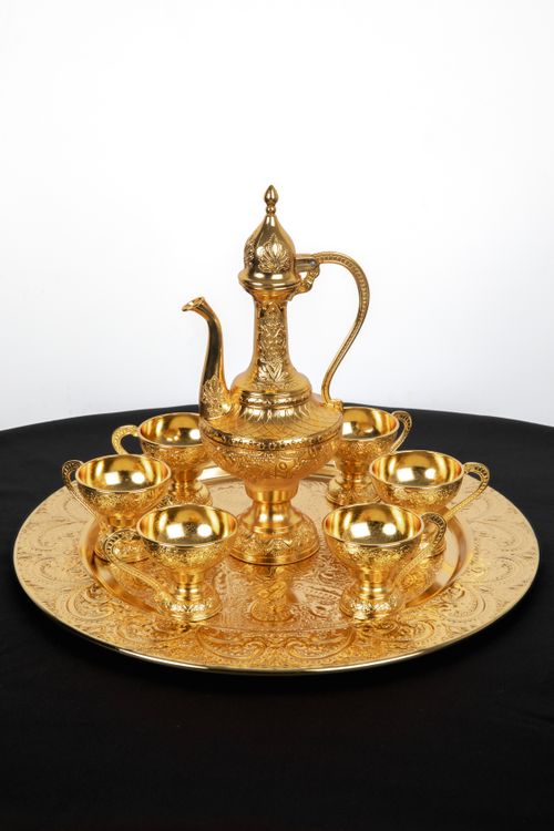 Free A luxurious golden Arabic coffee set featuring a traditional dallah with six cups on an intricately designed tray. Stock Photo