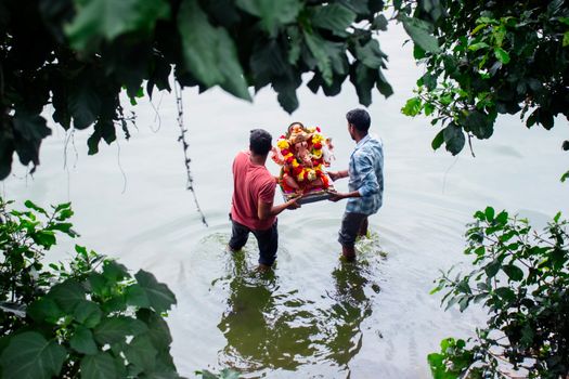 Two men perform a traditional Ganesh idol immersion in a river, surrounded by lush greenery.