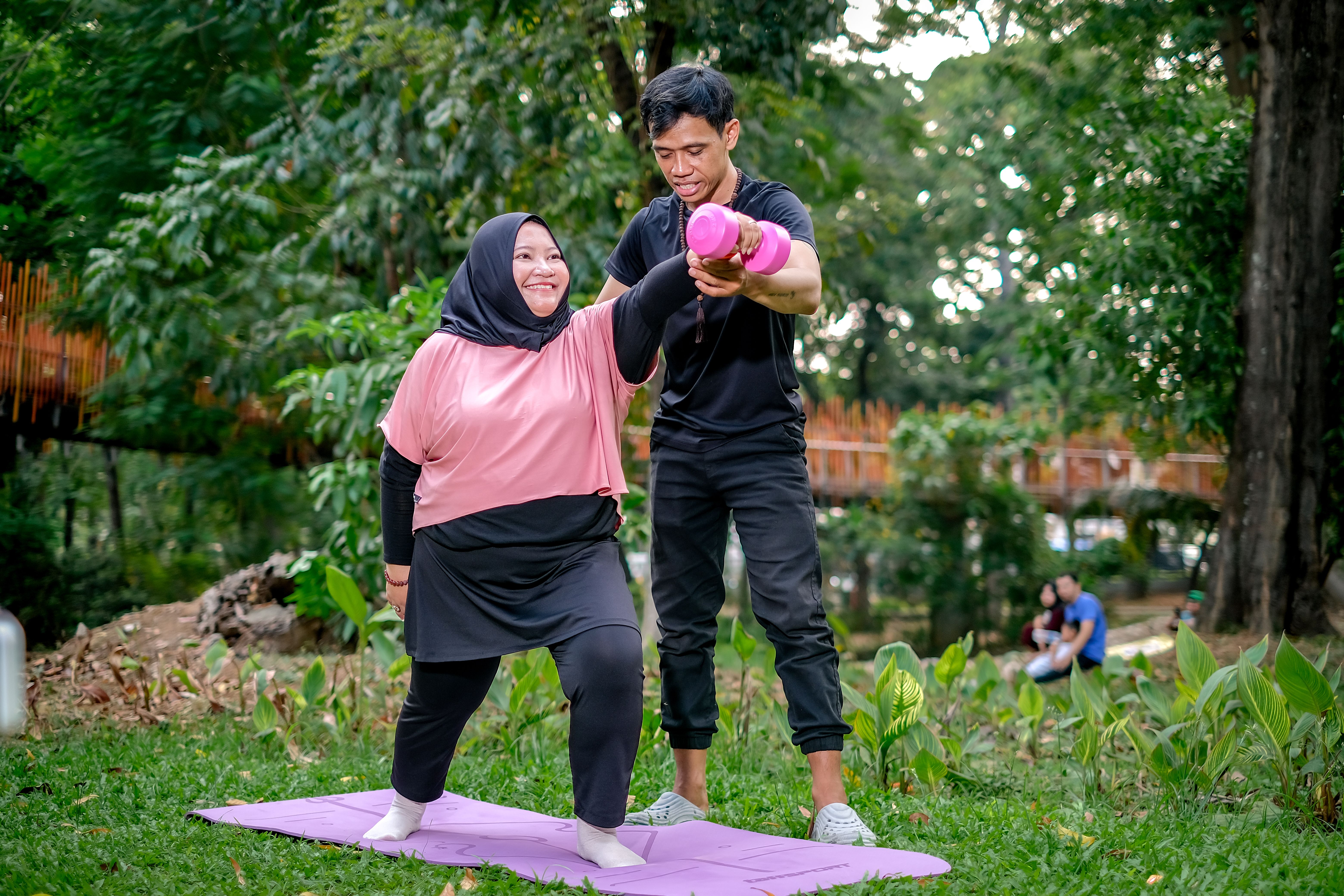 A personal trainer assists a woman during a yoga session in a lush Jakarta park.