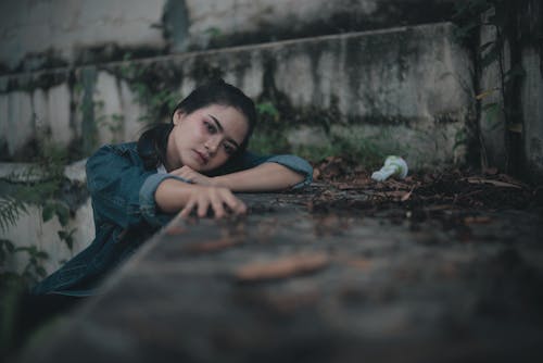 Sad pensive ethnic female teenager in jeans jacket looking at camera while sitting alone near old stone wall in park