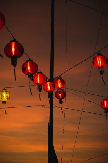 From below of colorful sphere shaped lanterns hanging on wires near wooden stick while illuminating street in evening under cloudy sky at sundown