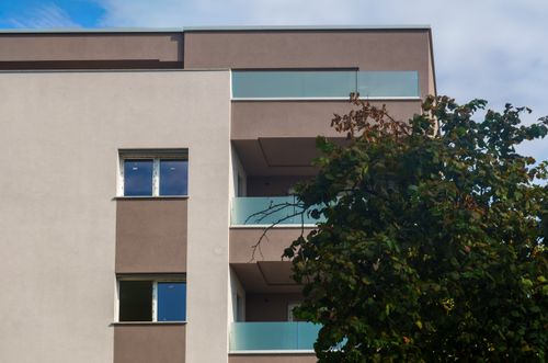 Free A modern apartment exterior with balconies and a tree in the foreground, under a blue sky. Stock Photo