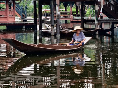 Elderly man in a wooden boat navigates a tranquil Asian canal with traditional architecture.