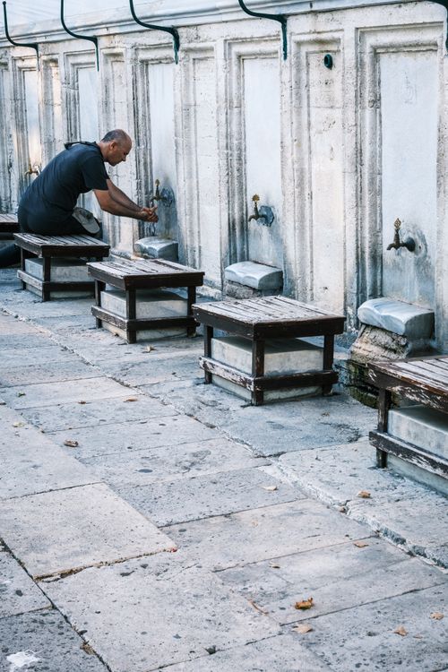 Free A man performs ablution at an outdoor water fountain with wooden benches. Stock Photo