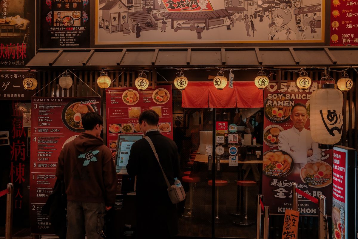 Two customers using a touchscreen ticket machine at the entrance of a ramen shop