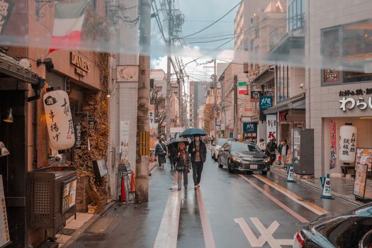 A lively street in Japan with pedestrians and shops under rainy weather.