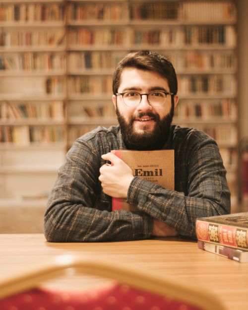 Free A bearded man sits in a library smiling, holding a book. Cozy and intellectual atmosphere. Stock Photo