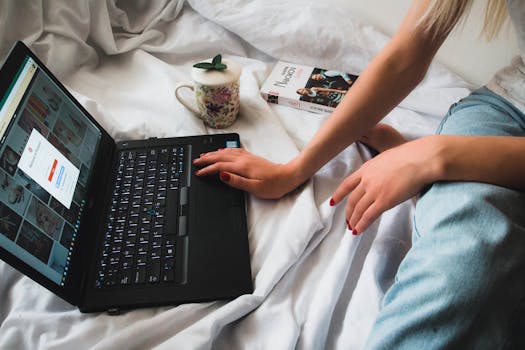 A person uses a laptop while resting on a bed, depicting a cozy work-from-home setup.