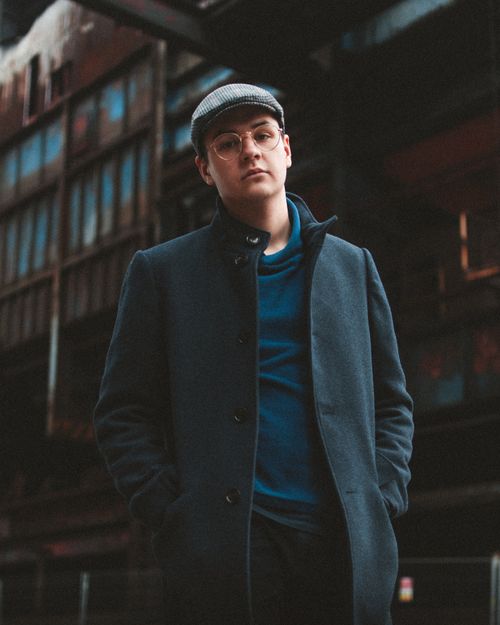 Free Low angle of modern young male in eyeglasses wearing classy coat and cap standing against blurred urban building Stock Photo