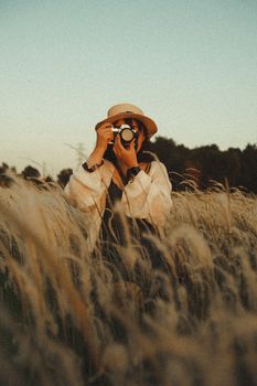 Anonymous woman in casual clothes and straw hat taking photo on digital camera while standing in field with overgrown spikes behind forest under serene sky in evening in countryside