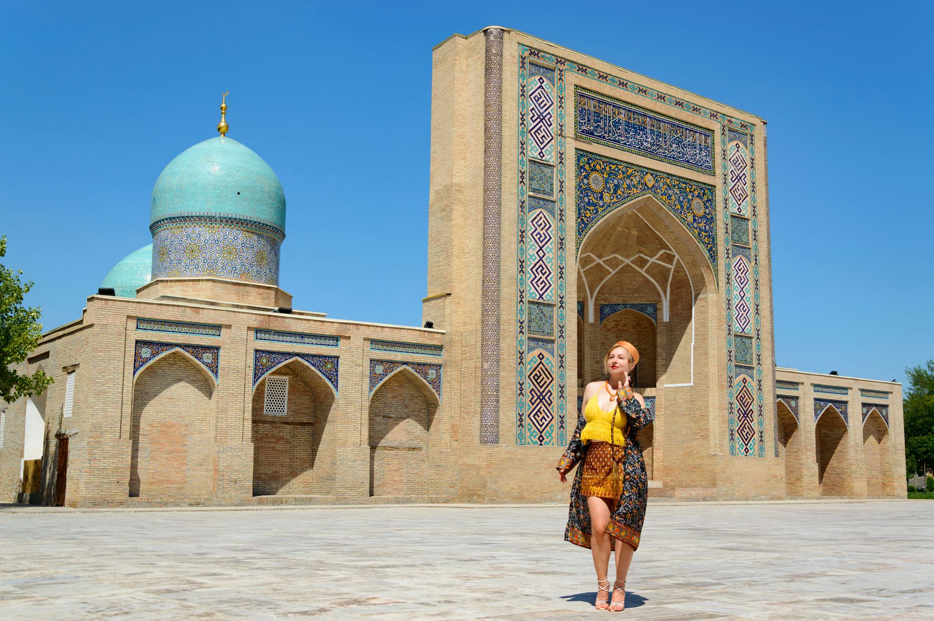 A woman poses in front of a historic Islamic architecture with intricate tiles and domes under a clear blue sky.