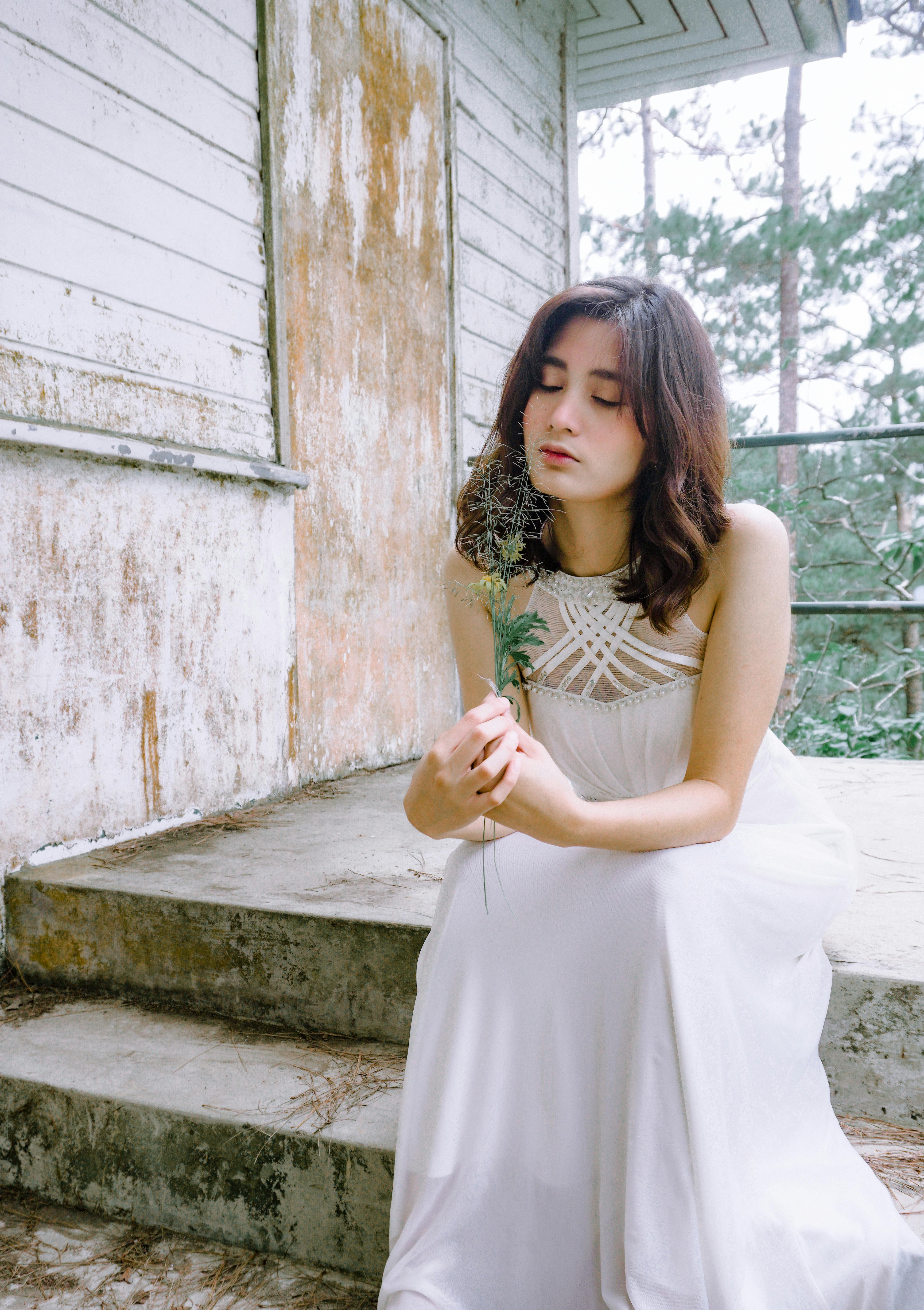 A serene portrait of a woman in a white dress holding a feather, seated on rustic steps outdoors.
