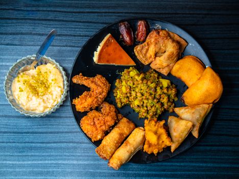 Delicious assortment of appetizers and desserts including samosas, shrimp, and flan on a dark table.