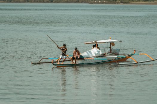 Three fishermen on a traditional boat navigate a calm lake under a sunny sky, using a wooden pole.