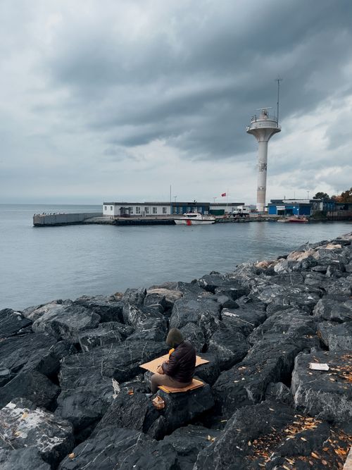 Free Person sitting by seaside near a lighthouse under cloudy skies. Stock Photo