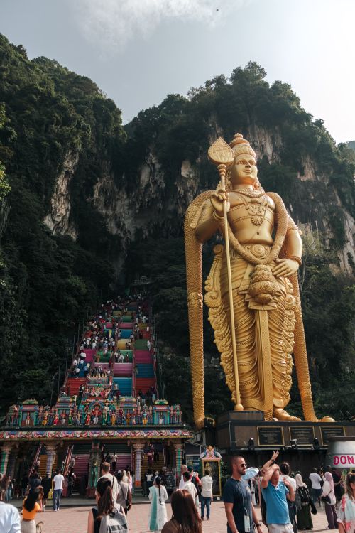 Free Golden Lord Murugan statue at Batu Caves, vibrant visitors ascend the colorful stairs. Stock Photo