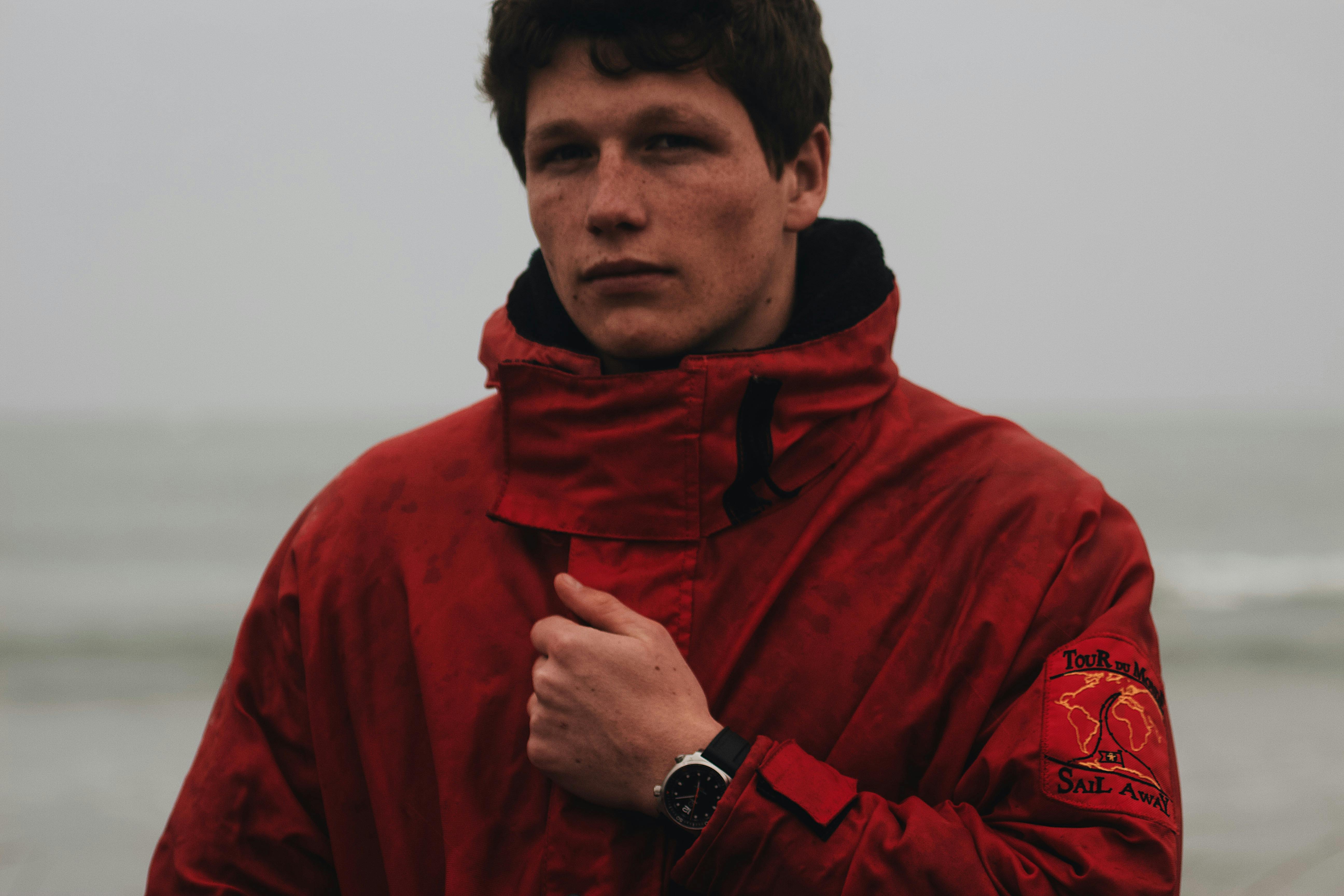 A man in a red jacket stands by the sea, exuding a thoughtful aura.