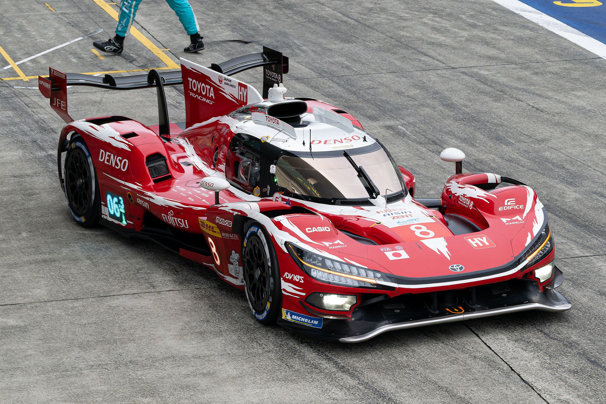 White and red race car in a pit lane at Fuji Speedway with team members nearby.