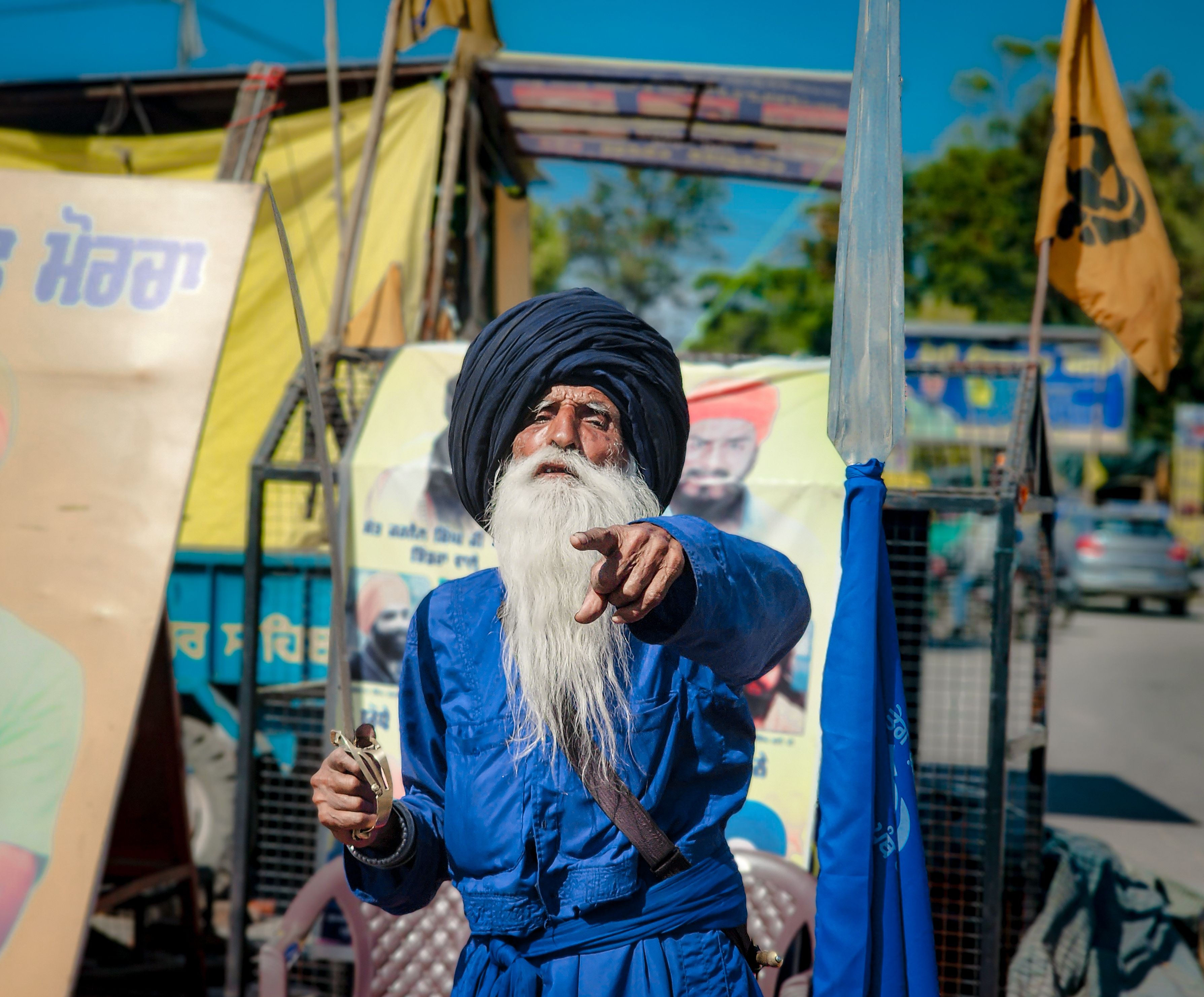 Elderly Sikh man in blue turban pointing during an outdoor gathering in Sahibzada Ajit Singh Nagar, India.