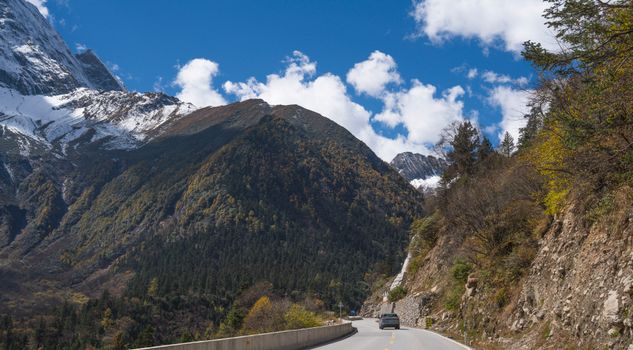 A car travels along a scenic road with Siguniang Mountain in the background under a clear blue sky.