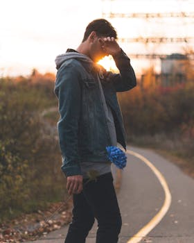 A contemplative man holding a blue flower walks on a path during sunset, depicting a serene and introspective moment.