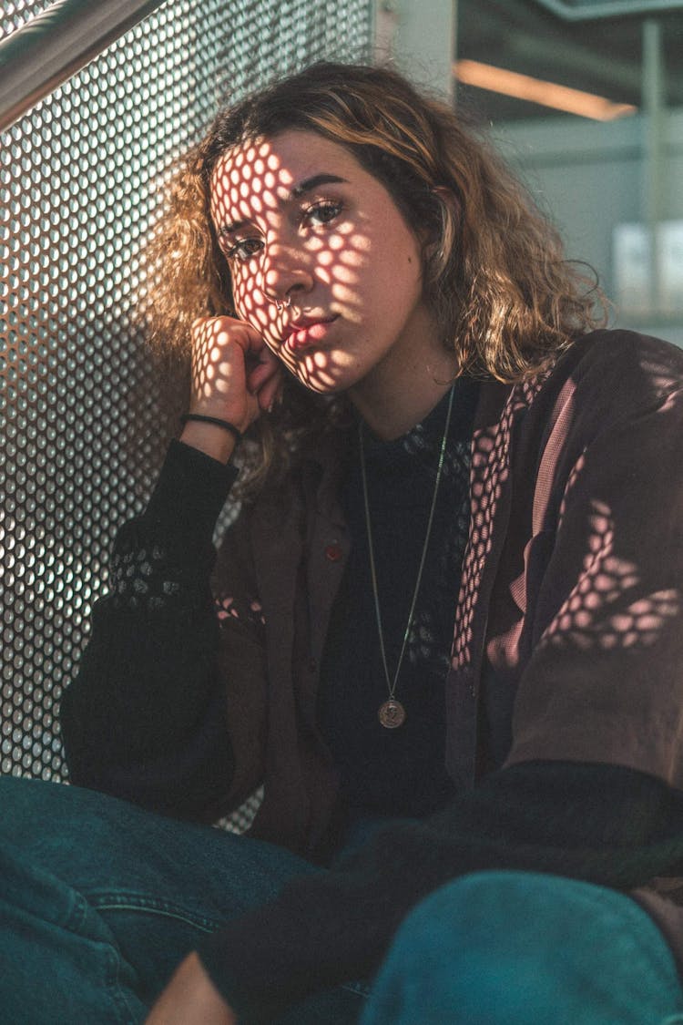 Dreamy Woman Sitting Near Grid Fence In Sunlight Indoors