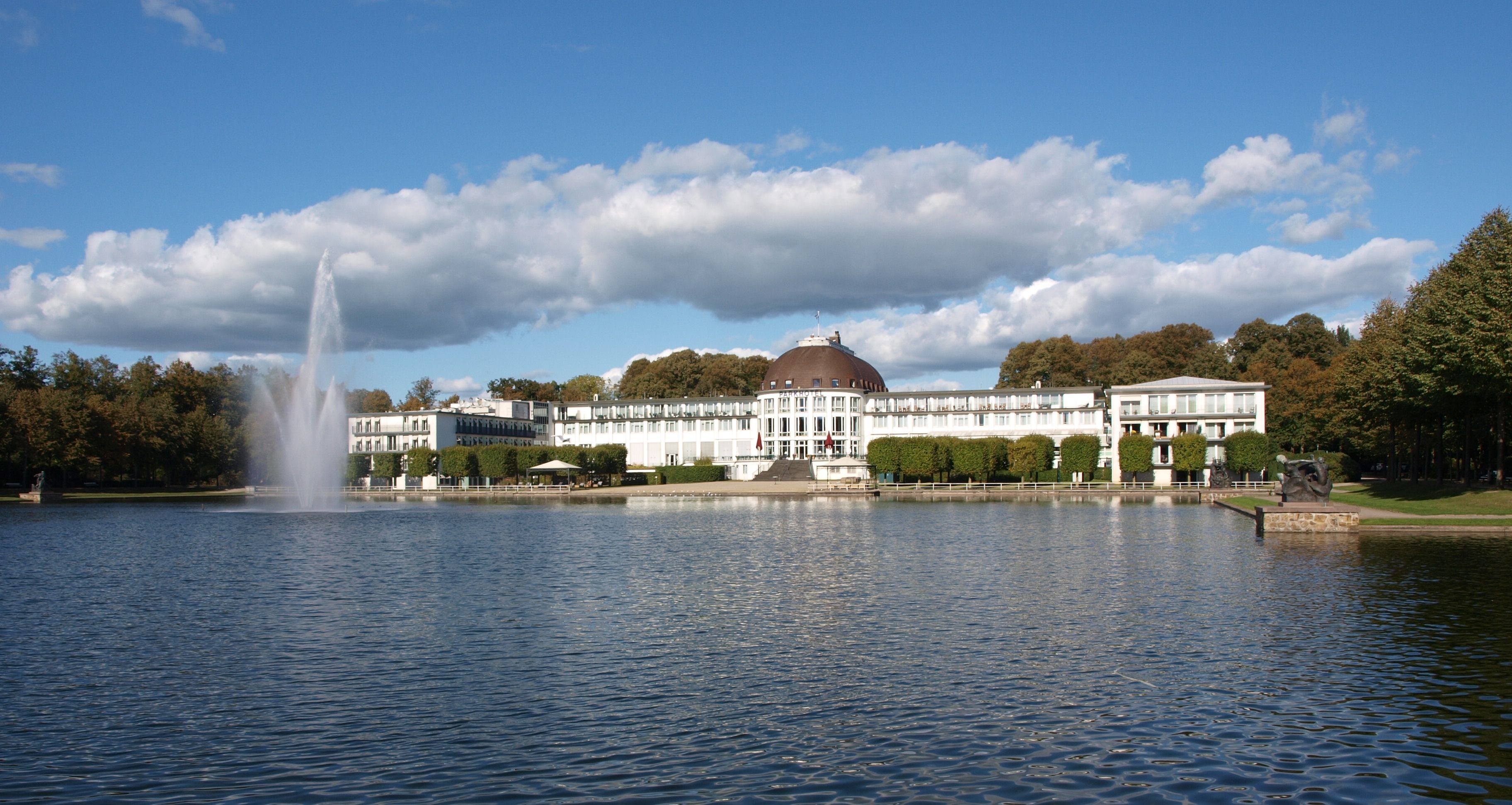 Scenic view of the Park Hotel Bremen with a reflecting lake and fountain in a tranquil park setting.