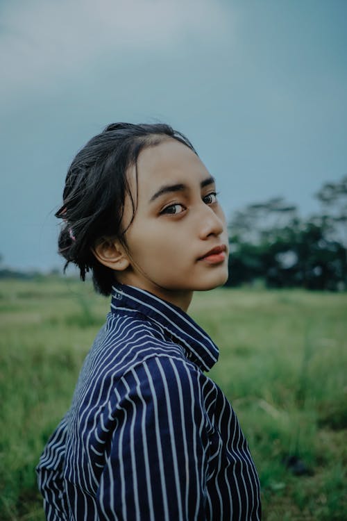 Side view of young pensive ethnic female in striped shirt looking away while standing on grass meadow behind growing trees under serene sky in daylight