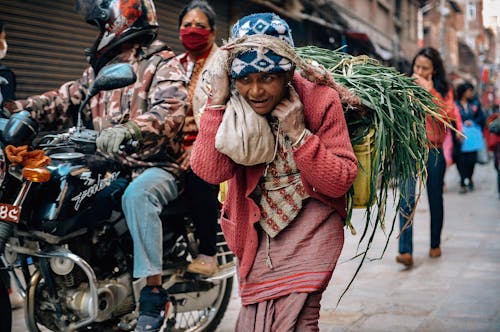 Mature ethnic female in national clothes carrying bunch of green herbs tied with rope while strolling on concrete pavement near couple on motorbike and houses in town and looking away