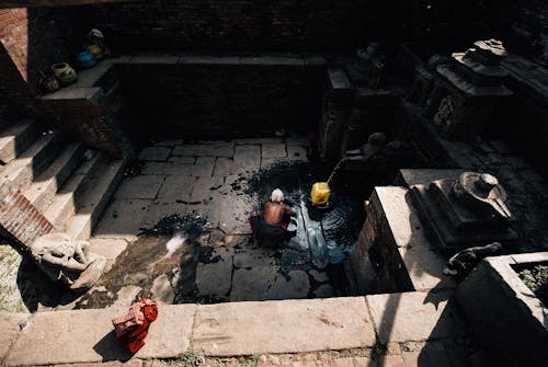 From above back view of anonymous elderly shirtless male squatting on dirty cement floor near plastic yellow container under water flow from well near stairs in old building in sunlight