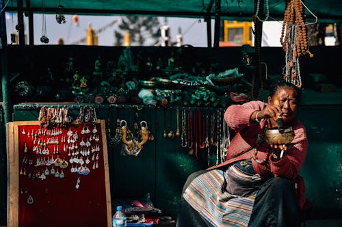 Serious mature overweight ethnic female vendor sitting with pounder and pestle near stands with collection of accessories in city and looking away