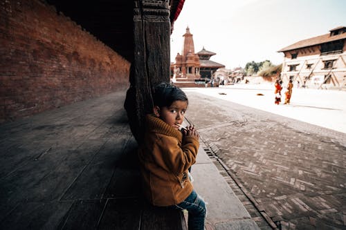 From above side view of adorable little ethnic kid sitting with hands clasped on wooden platform of building with brick wall while looking at camera and biting stick