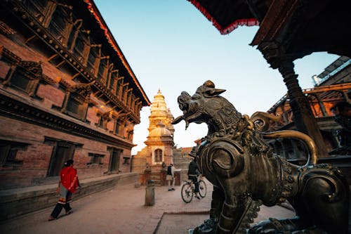 From below of aged ornamental statue of animal in front of stone church facade with cupola and unrecognizable tourists walking on pavement under serene sky in evening