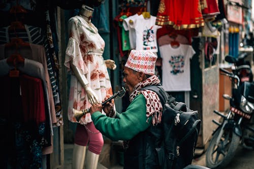 Side view of anonymous mature ethnic male artist with backpack playing musical instrument while standing in front of local clothing shop on city street in daylight near parked motorbike