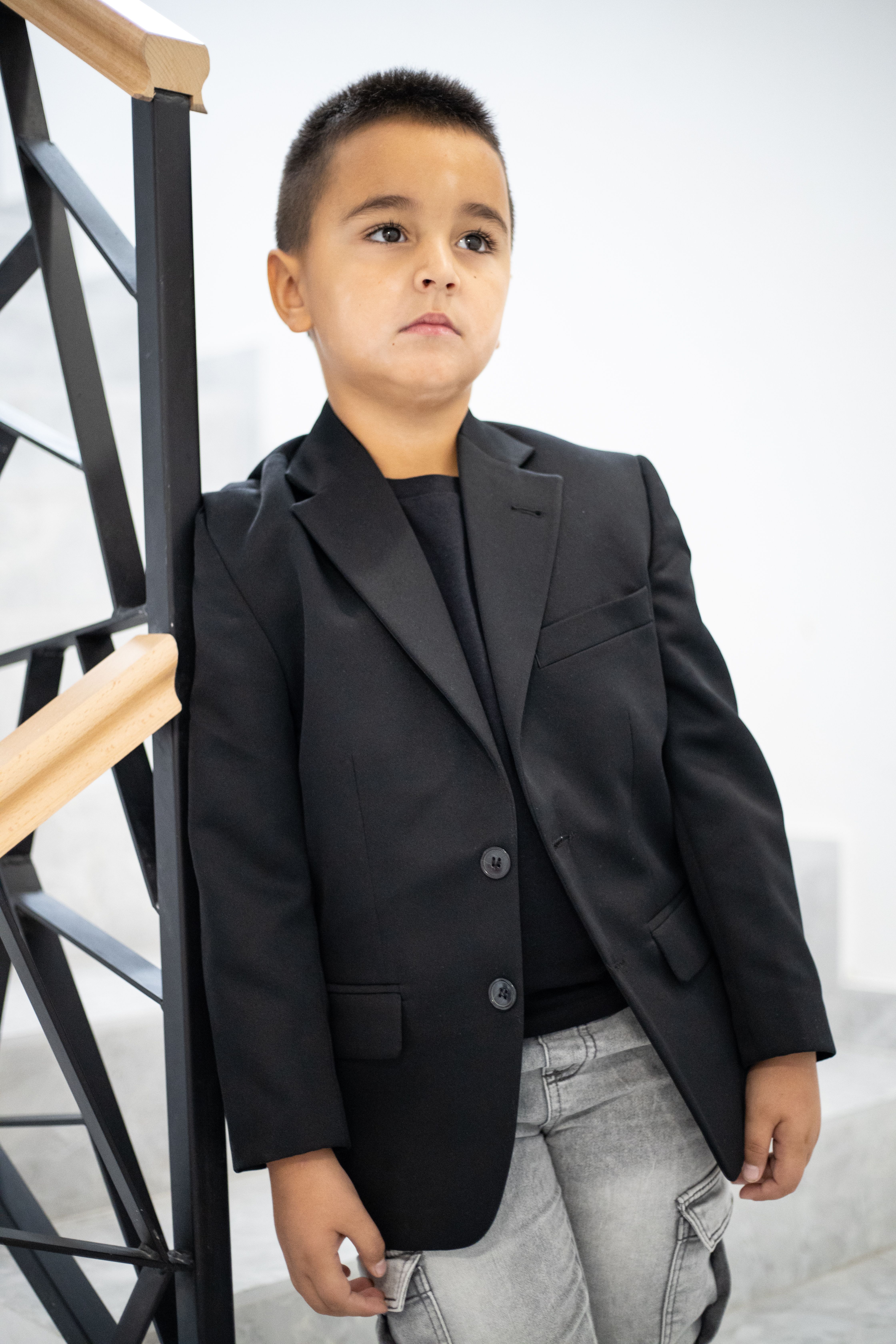 Fotomural Retrato de un niño pequeño con una chaqueta negra apoyado en una escalera moderna en el interior de una vivienda.