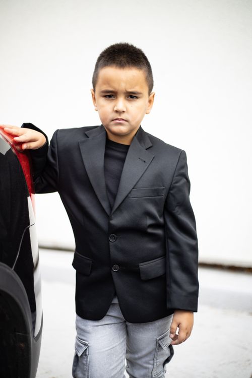 Free A young boy in a stylish black suit stands confidently beside a vehicle, exuding a mature and serious demeanor. Stock Photo