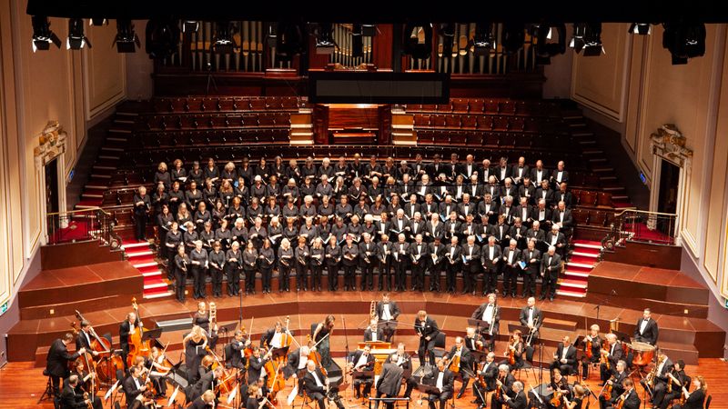 Großes Sinfonieorchester mit Streichern im Vordergrund und einem Chor in schwarzer Kleidung auf den Rängen eines Konzertsaals mit Orgel