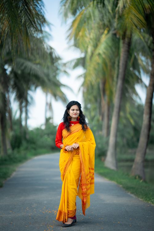 Free South Asian woman in vibrant yellow saree walking on a road lined with lush palm trees, symbolizing cultural elegance. Stock Photo