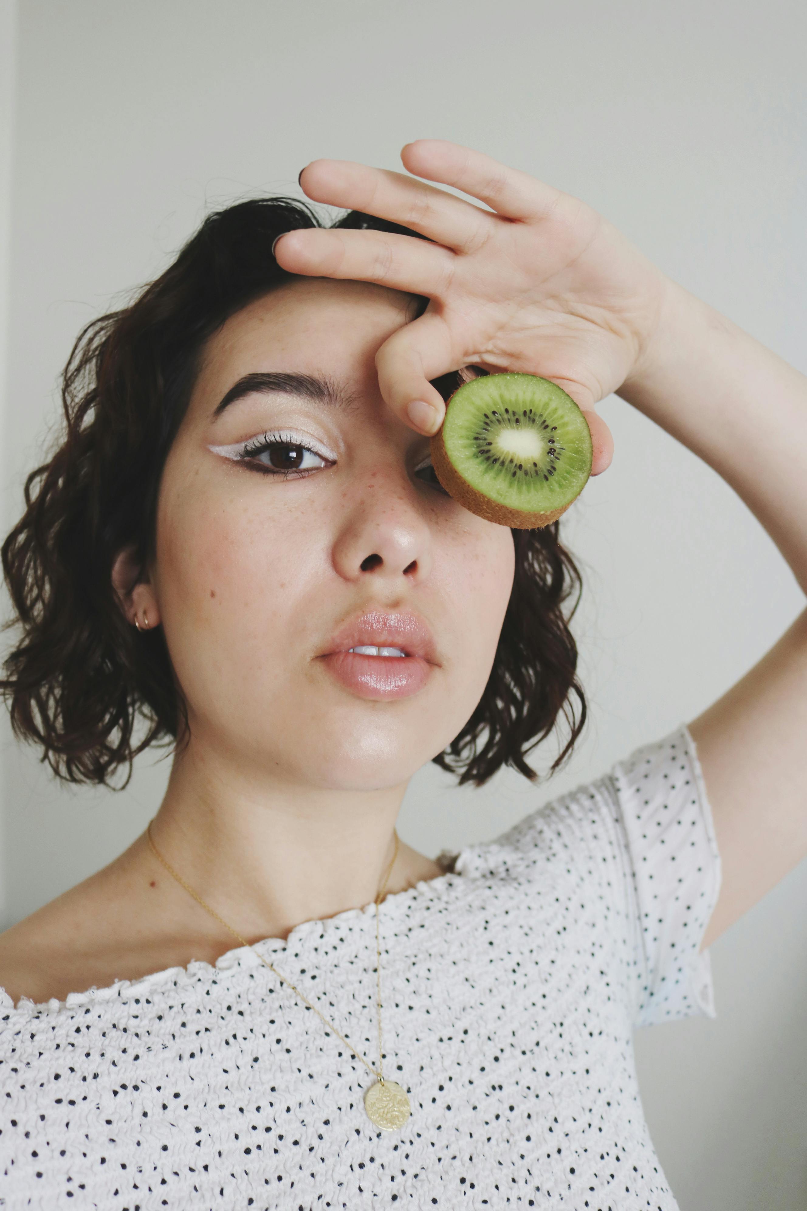 Artistic portrait of a young woman holding a kiwi fruit over one eye, with creative makeup.