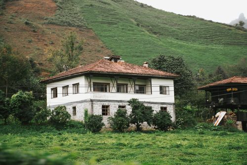 Free Two-story country house surrounded by lush greenery and rolling hills, creating a serene rural scene. Stock Photo
