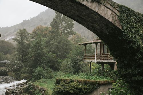 Free A serene riverside cabin nestled under a rustic stone bridge surrounded by lush greenery. Stock Photo
