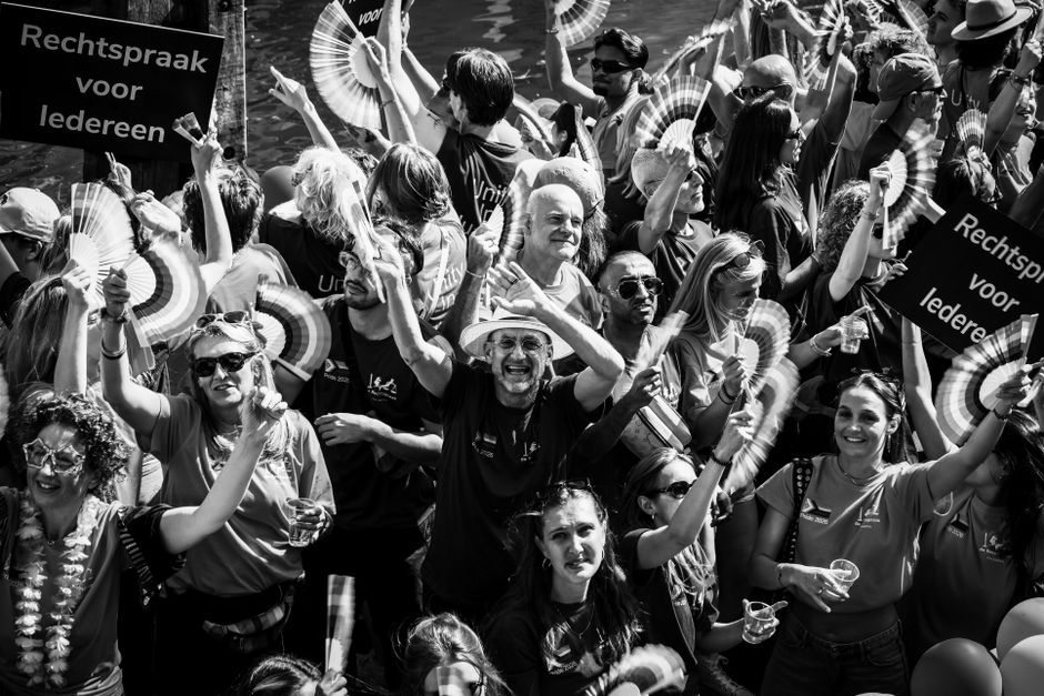 Black and white photo of a lively crowd celebrating at an Amsterdam parade, holding fans and signs.