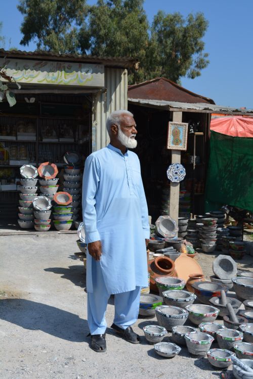 Free An elderly man in traditional attire stands outside a Pakistani pottery shop surrounded by colorful clay pots. Stock Photo