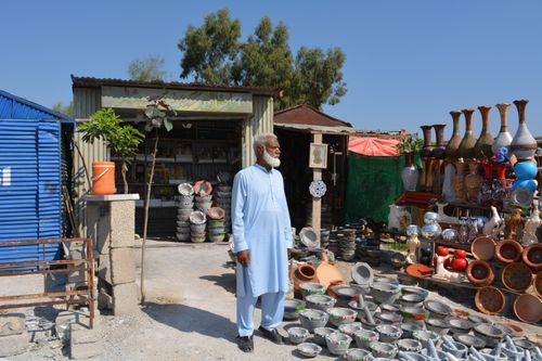 Free Pakistani artisan standing outside a shop displaying traditional pottery and crafts. Stock Photo