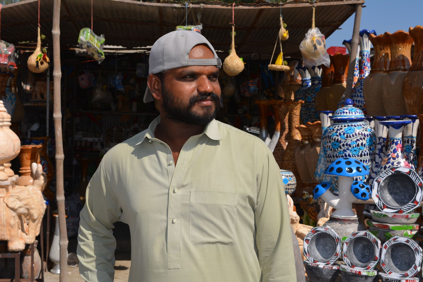 A young man stands in front of a Pakistani handicraft shop displaying colorful pottery and sculptures.