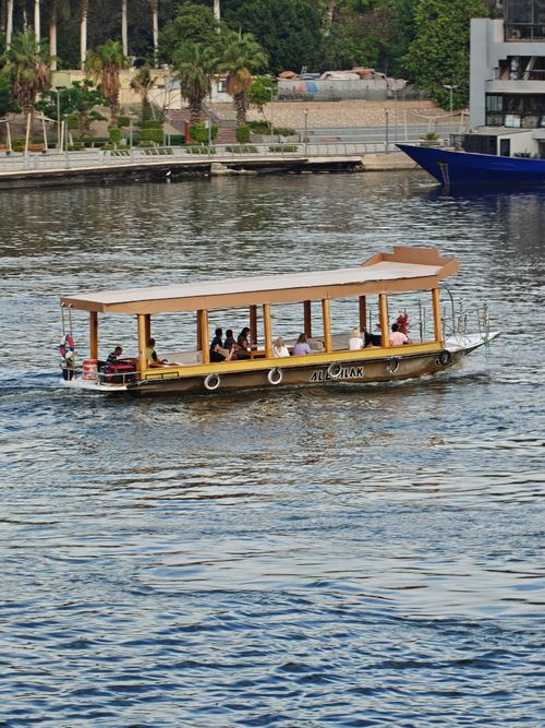 Free A covered boat carrying passengers on a vibrant city river, surrounded by greenery and buildings. Stock Photo